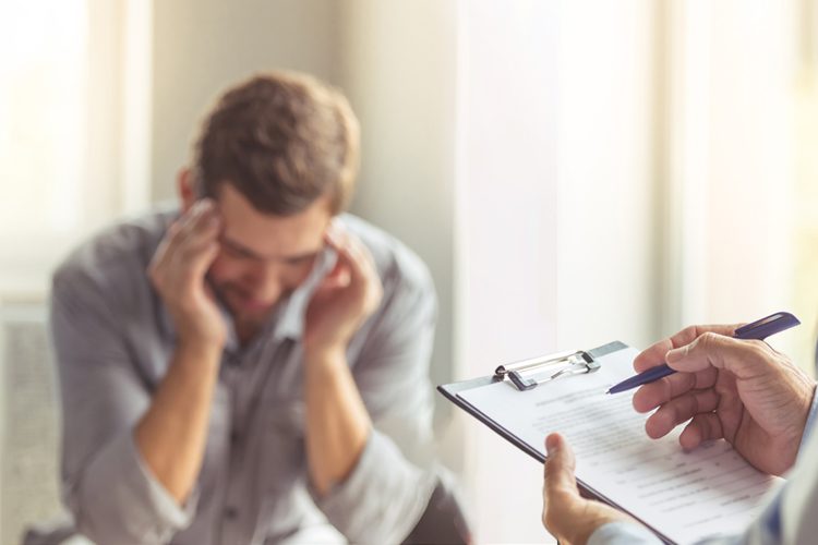 Mental Health Specialized Services: Man holding his temples, leaned forward in a as a counselor makes notes